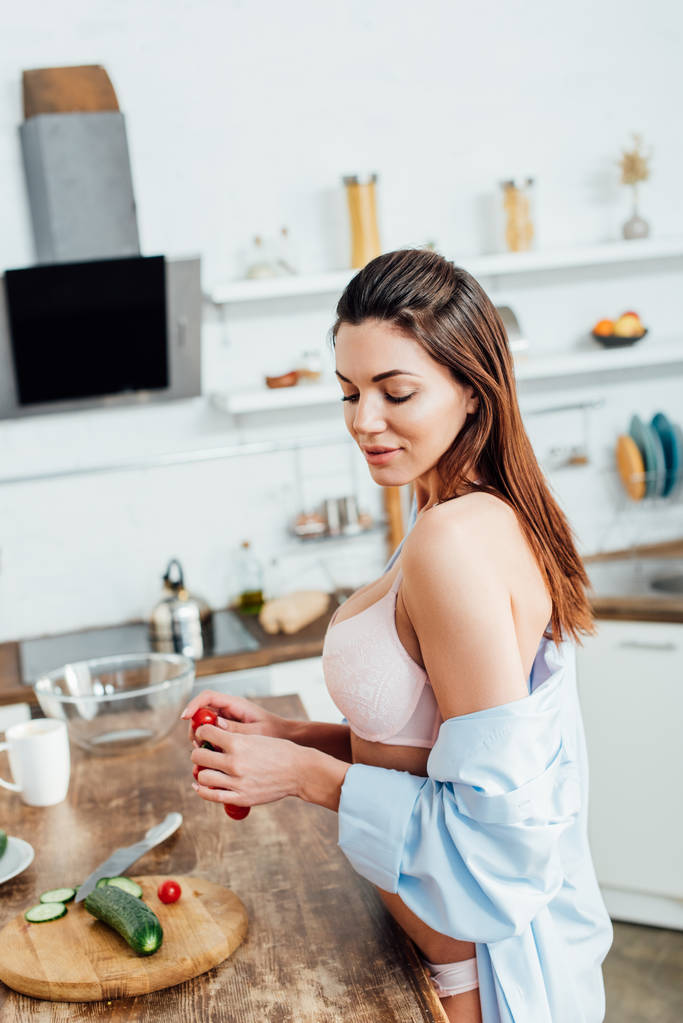 Smyslová dívka v podprsence a košili, držící v kuchyni třešňová rajčata - Fotografie, Obrázek