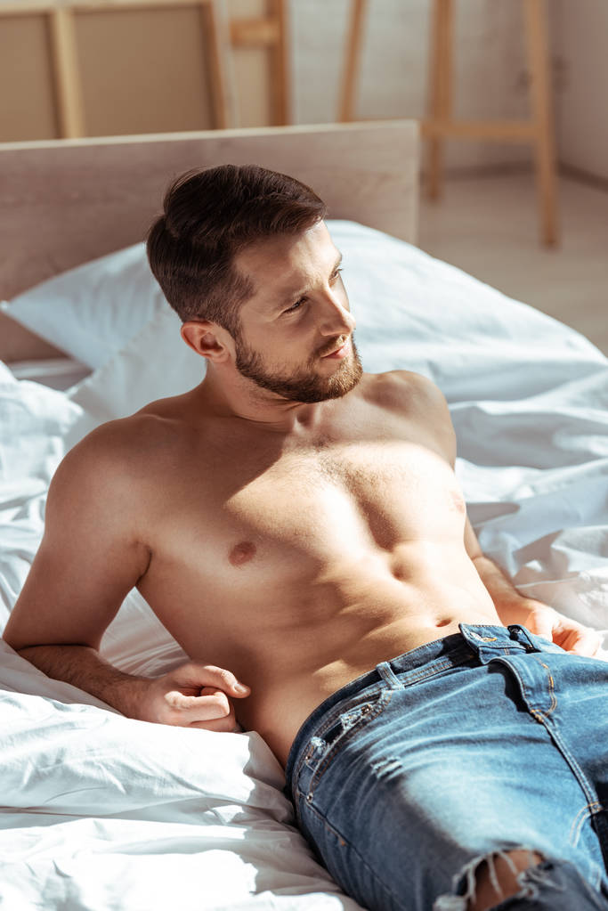 hombre guapo y sin camisa acostado en la cama y mirando hacia otro lado en el dormitorio
  - Foto, Imagen