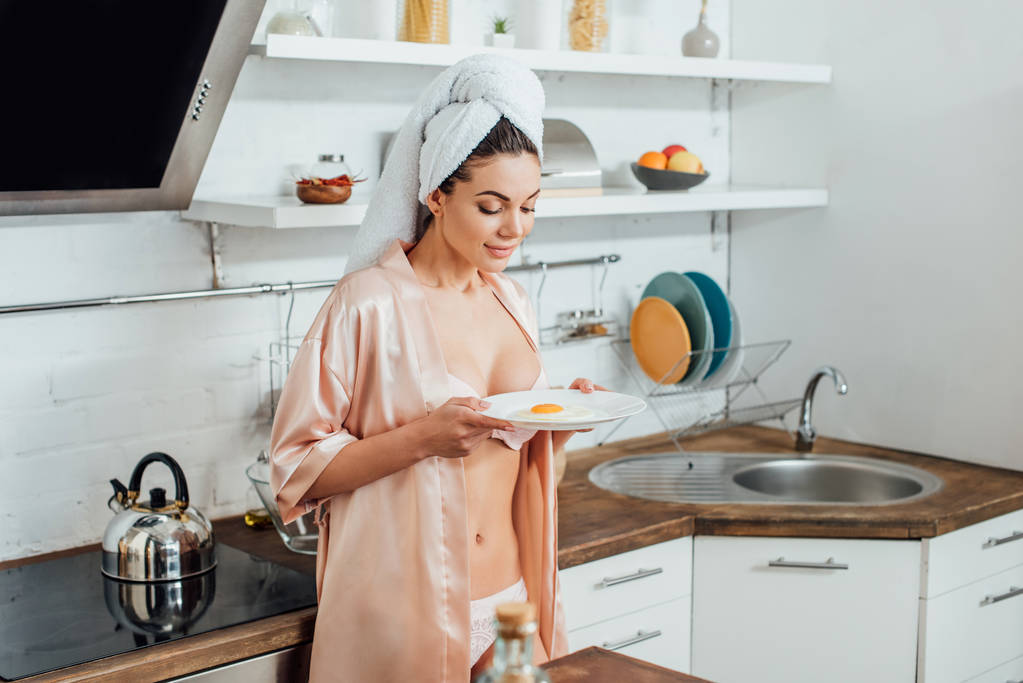 Sexy woman in housecoat holding plate with fried egg in kitchen - Photo, Image