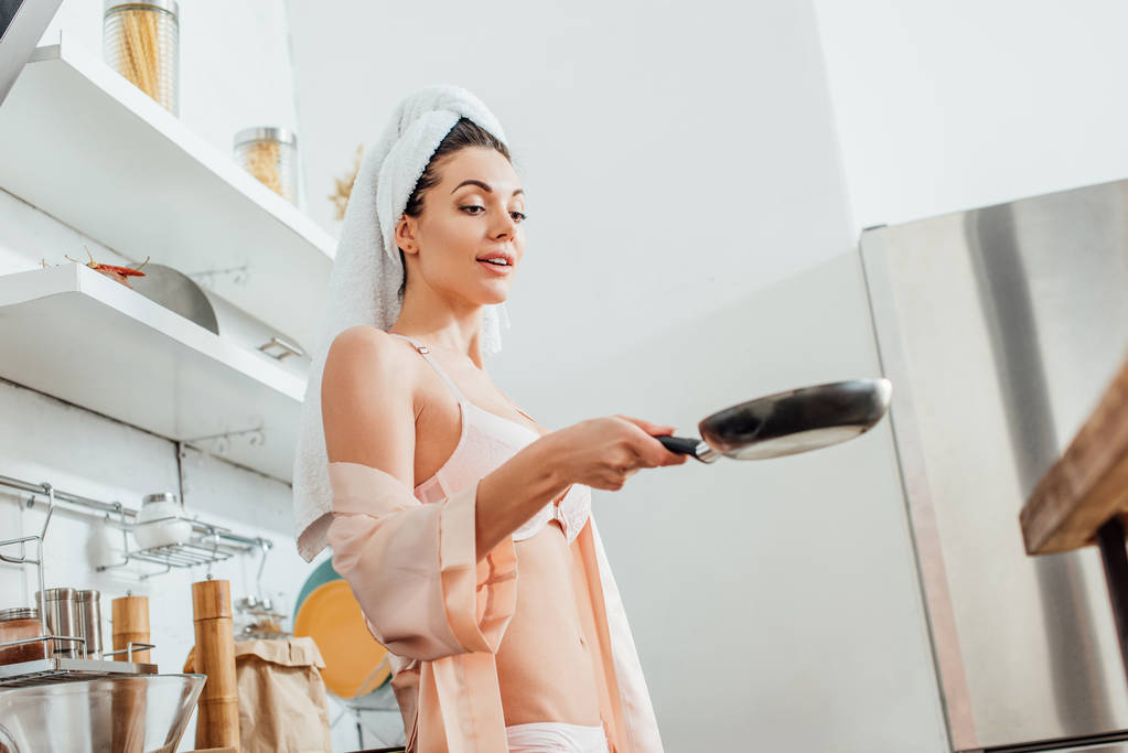 Niski kąt widzenia seksownej dziewczyny w housecoat z ręcznikiem na głowie trzymając patelnię w kuchni - Zdjęcie, obraz