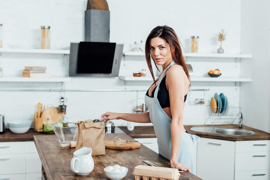 Zúčastněná žena ve spodním prádle a zástěře stojící blízko dřevěného stolu v kuchyni - Fotografie, Obrázek