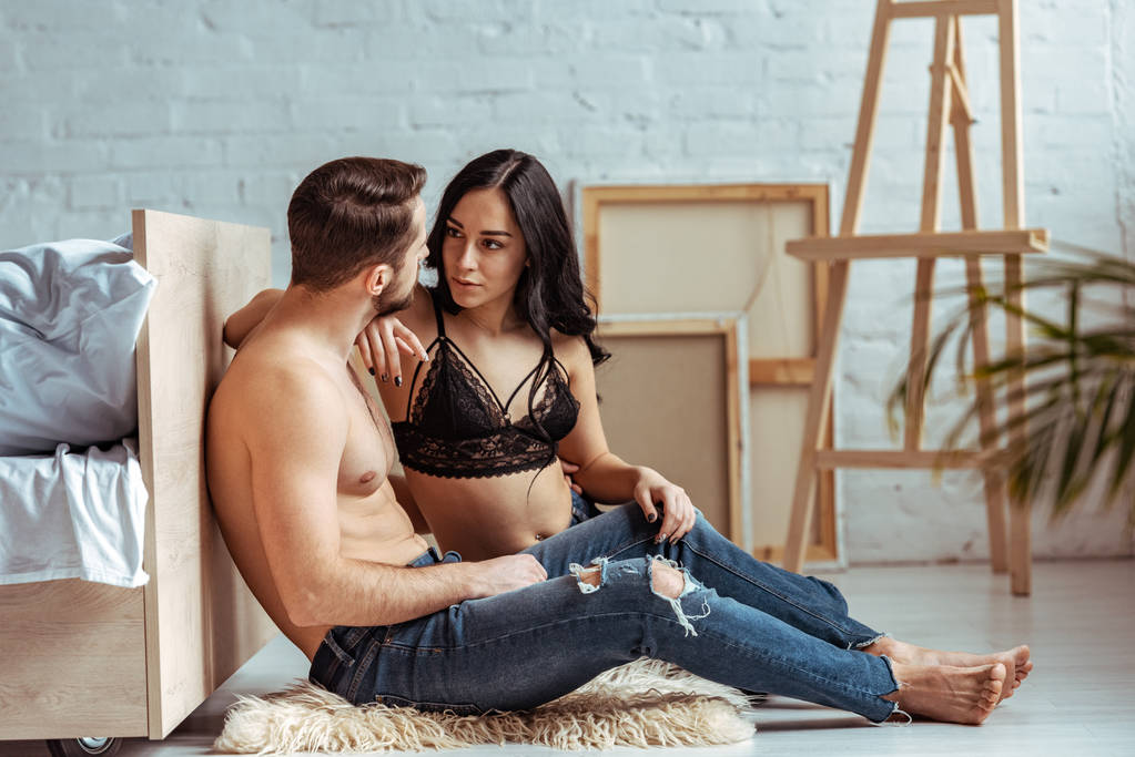 hombre guapo y musculoso sentado en la alfombra y abrazándose con hermosa mujer en sujetador de encaje en el dormitorio
  - Foto, imagen