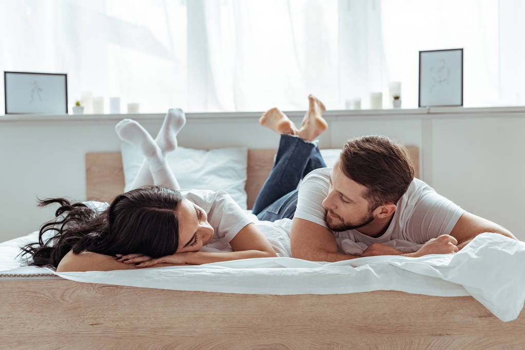 vriendje in jeans en vrouw in korte broek en t-shirt liggend op bed en kijken elkaar in slaapkamer  - Foto, afbeelding