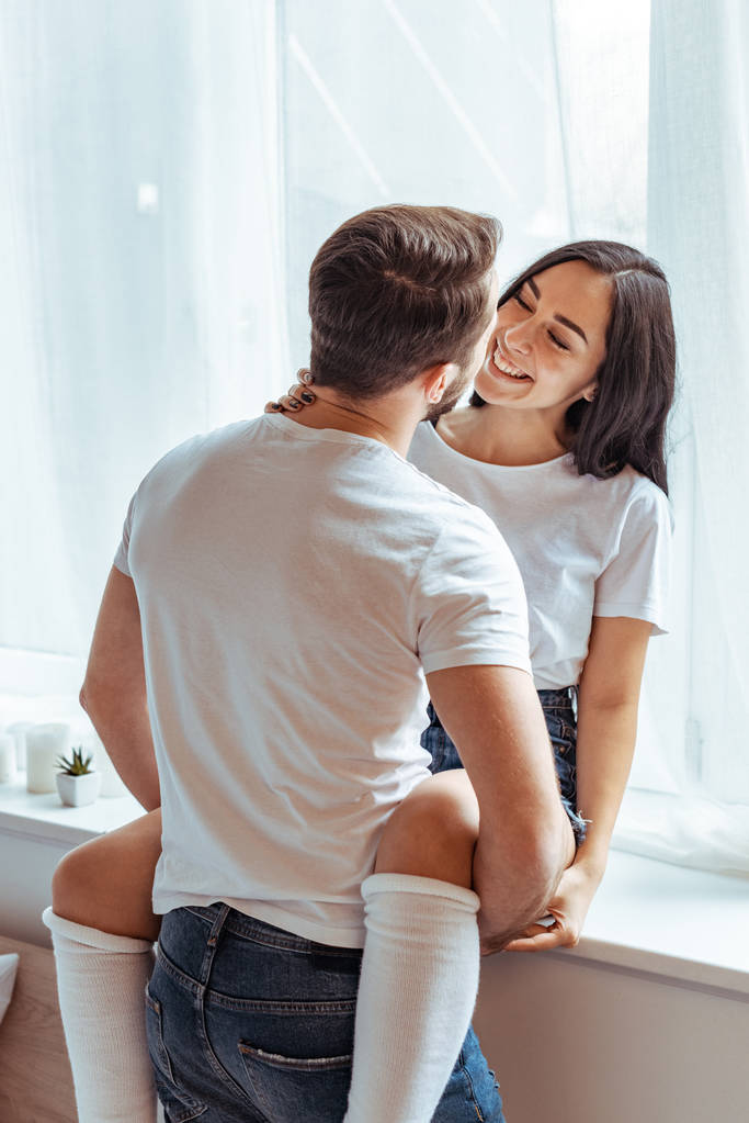 man in het witte t-shirt knuffelen mooie en lachende vrouw in slaapkamer  - Foto, afbeelding