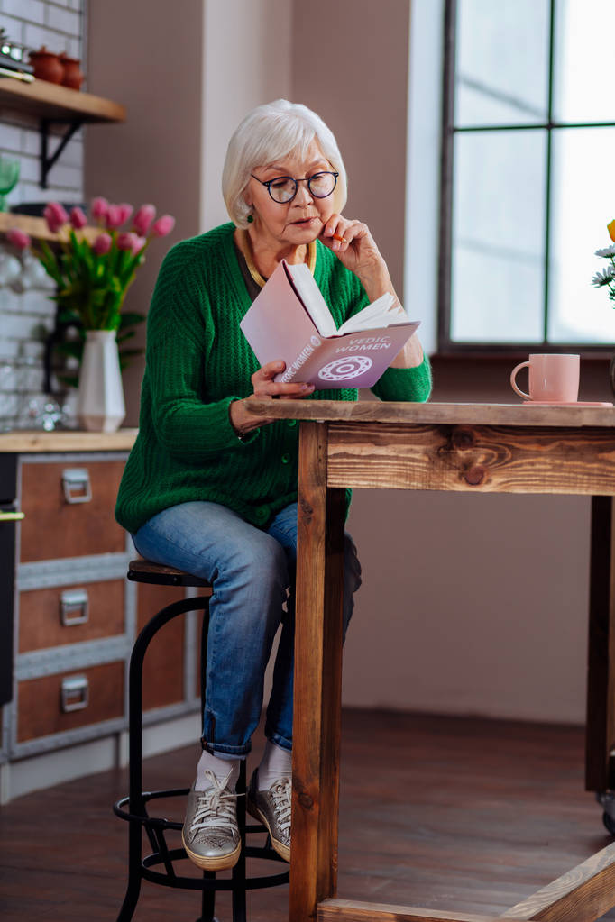 Стареющая женщина поглощена книгой, сидя на меблированной кухне
 - Фото, изображение