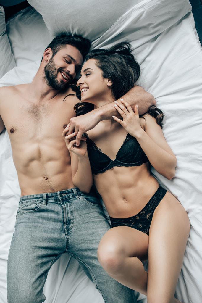 Blick von oben auf einen glücklichen Mann ohne Hemd, der eine lächelnde Frau in Spitzenunterwäsche ansieht, während er auf dem Bett liegt - Foto, Bild