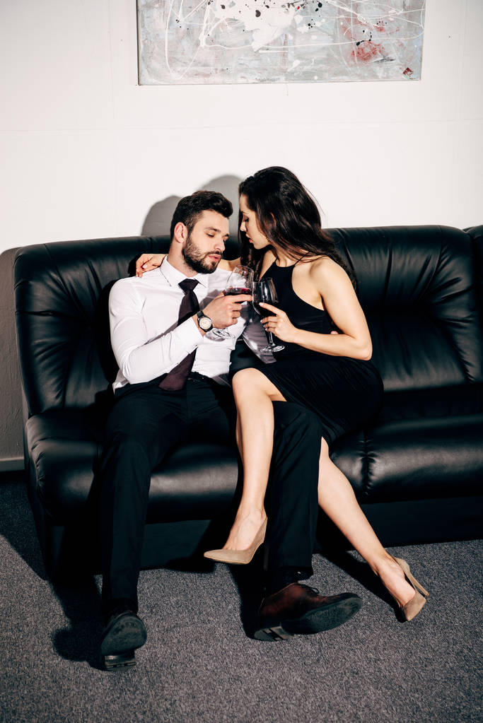 όμορφος άνδρας και όμορφη γυναίκα στο μαύρο φόρεμα κάθεται στον καναπέ και κρατώντας ποτήρια κρασί  - Φωτογραφία, εικόνα