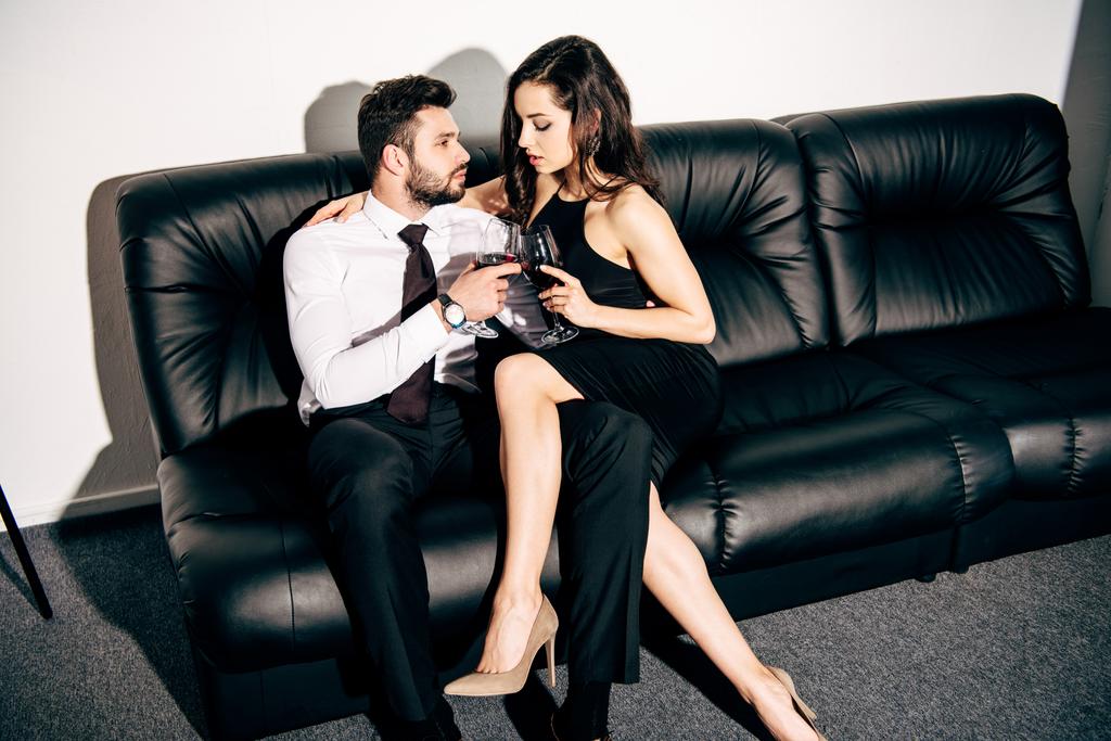 barbudo homem e mulher em vestido preto sentado no sofá e segurando copos de vinho
  - Foto, Imagem