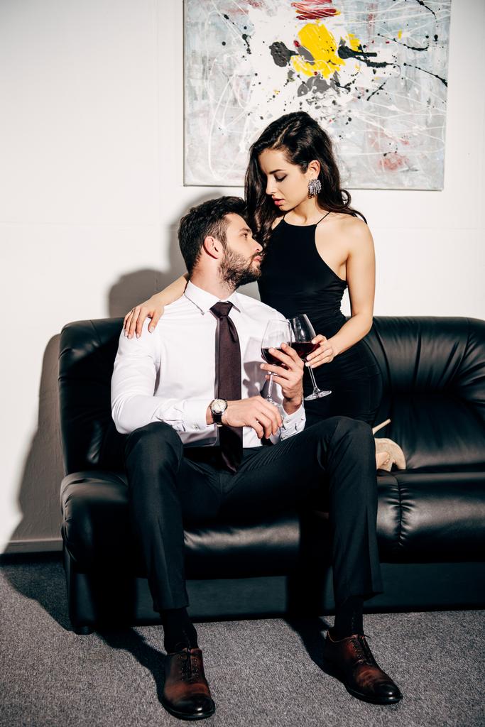  Brodaty mężczyzna ogląda dziewczyna w czarnej sukni siedzi na kanapie i trzymając kieliszek wina  - Zdjęcie, obraz