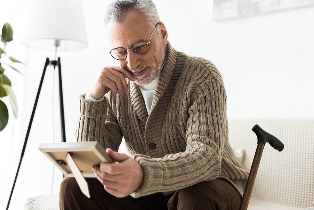Veselý bývalý muž drží fotorámeček zatímco sedí na pohovce nedaleko vycházkovou hůl - Fotografie, Obrázek