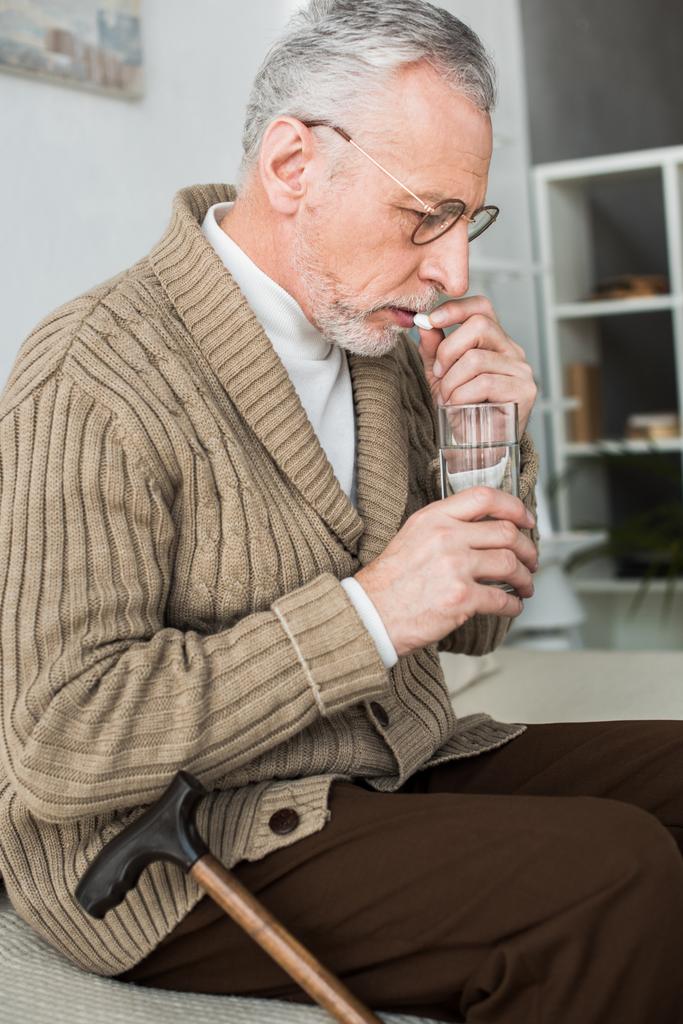 пенсіонер приймає таблетку, сидячи на дивані біля прогулянкової тростини
  - Фото, зображення
