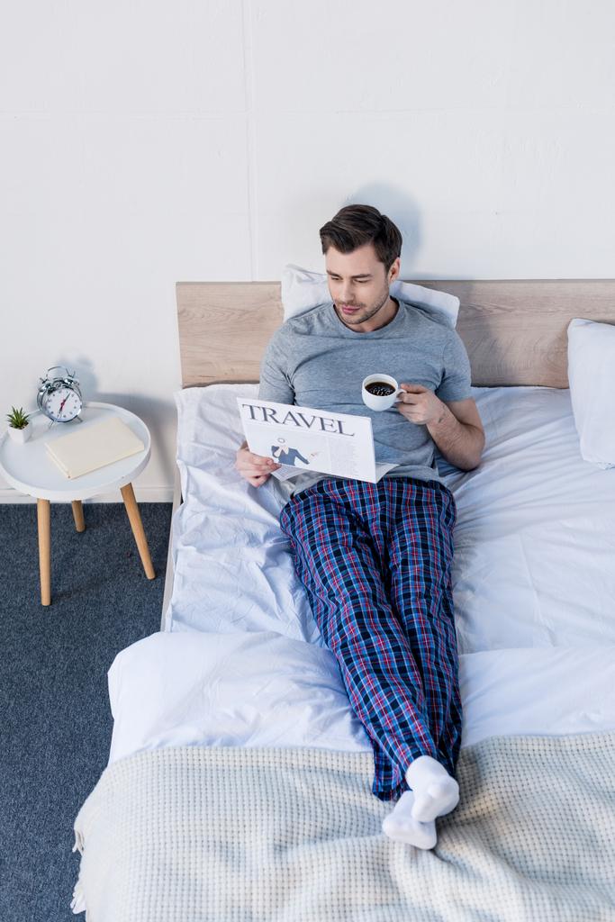 コーヒーと旅行新聞でベッドの上に休んでいるパジャマのハンサムな男 - 写真・画像