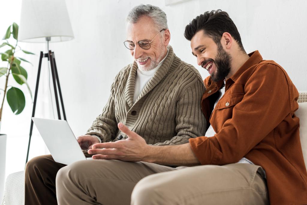 fröhlicher bärtiger Mann gestikuliert, während er mit dem älteren Vater sitzt und auf Laptop schaut  - Foto, Bild