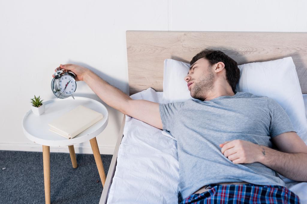 przystojny overslept mężczyzna trzyma budzik podczas układania na łóżku w domu - Zdjęcie, obraz
