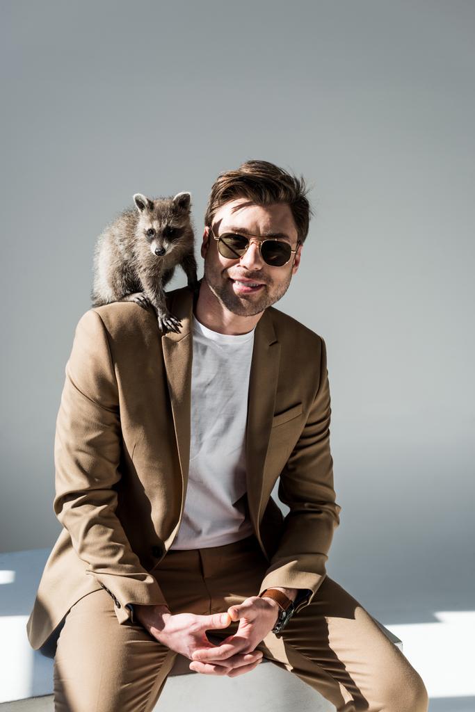 handsome man in sunglasses with cute raccoon on shoulder on grey - Photo, Image