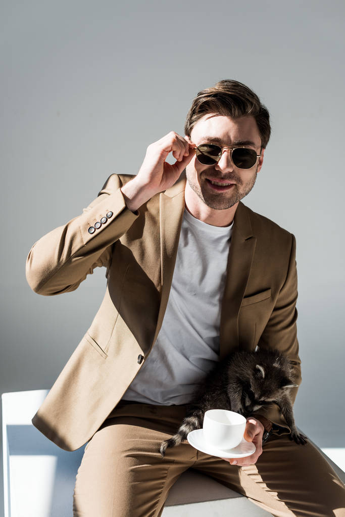 hombre guapo con mapache divertido y taza de café en vueltas mirando a la cámara en gris
 - Foto, imagen