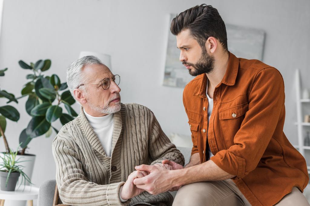sad senior father in glasses holding hands with handsome son at home - Photo, Image