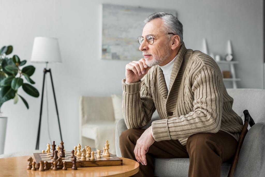 Nachdenklicher Rentner mit Brille, der daheim am Schachbrett sitzt - Foto, Bild