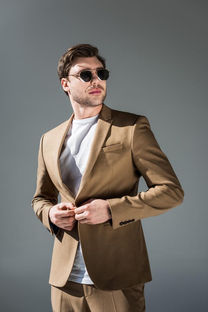 hombre de moda de confianza en suite beige y gafas de sol mirando hacia otro lado en gris
 - Foto, Imagen