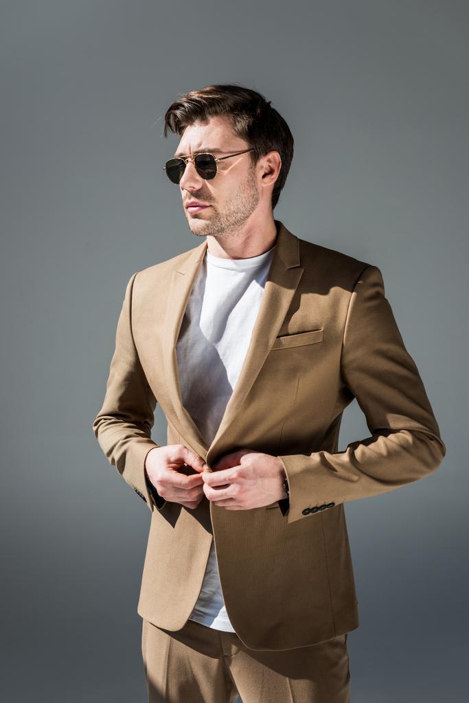 serieuze stijlvolle man in beige Suite en zonnebrillen op zoek weg op grijs - Foto, afbeelding