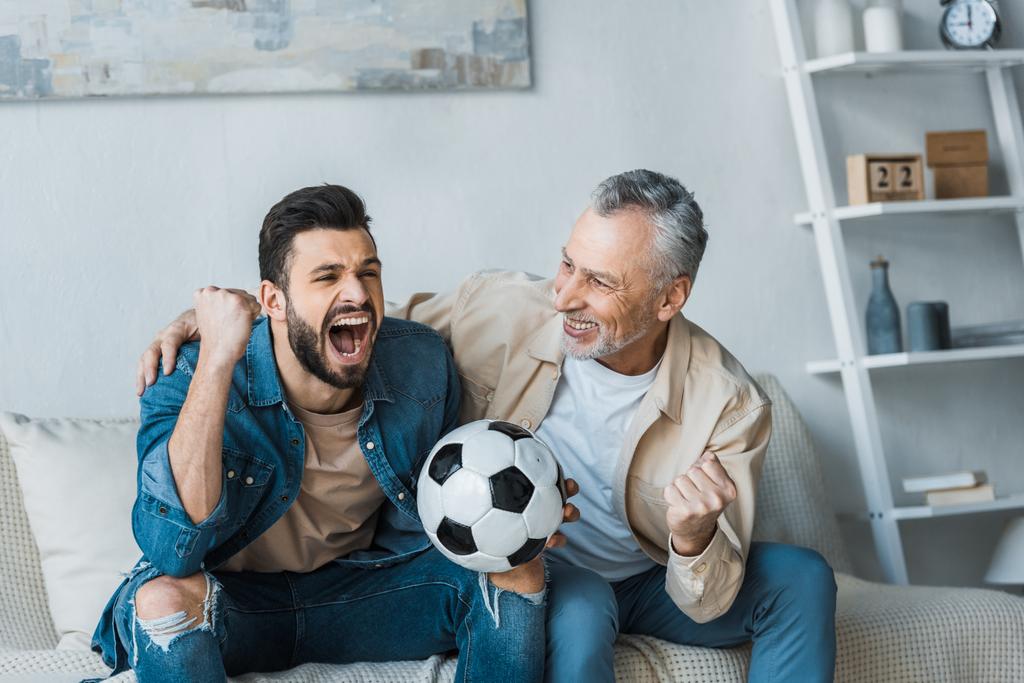 старший чоловік дивиться на щасливого сина підбадьорює під час перегляду чемпіонату і тримає футбол
  - Фото, зображення