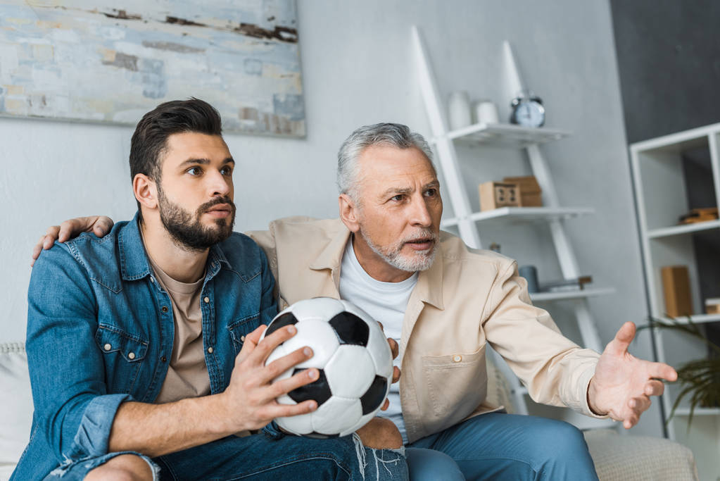 サッカーを保持しているハンサムな息子と選手権を見ながらジェスチャー引退した男  - 写真・画像