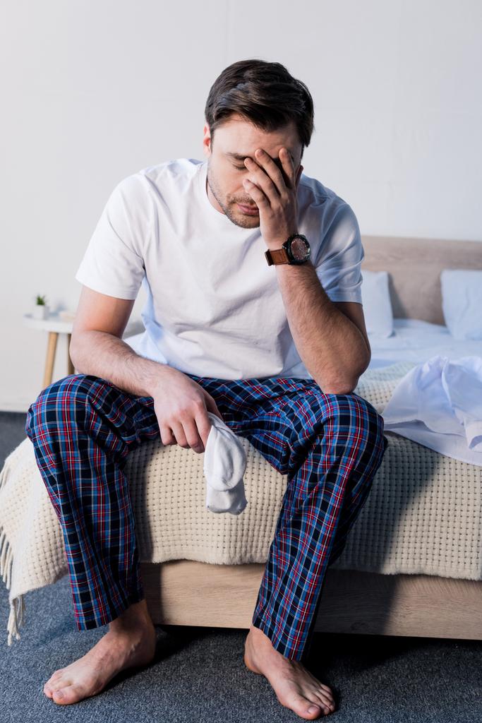 Красивый сонный мужчина сидит на постелях и держит белые носки
 - Фото, изображение