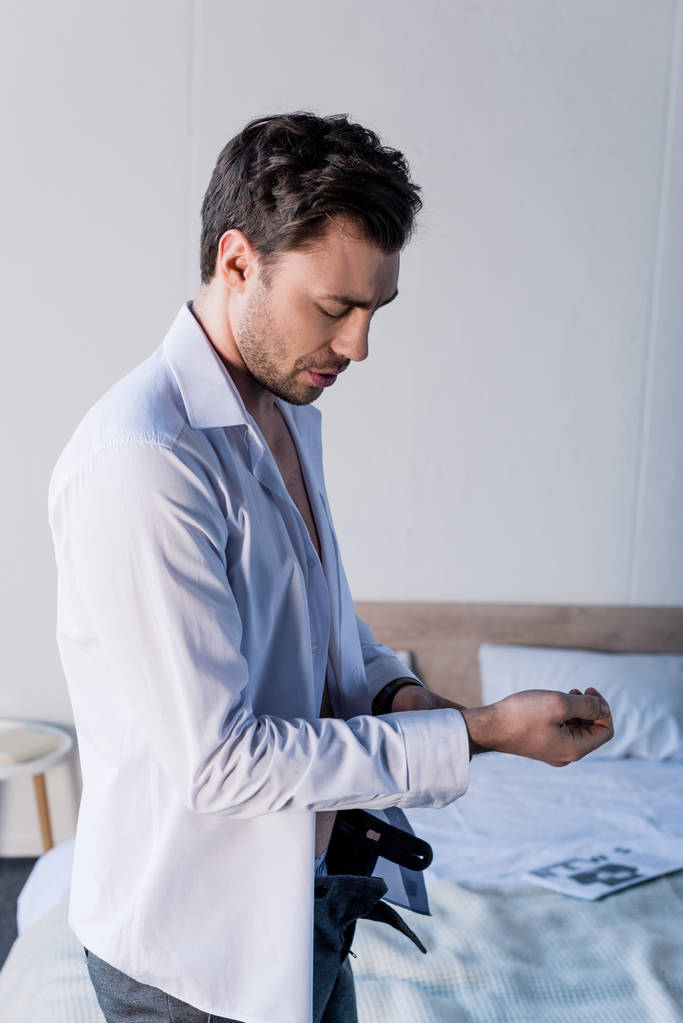 όμορφος νυσταγμένος άντρας ντύνει λευκό πουκάμισο ενώ στέκεται κοντά στο κρεβάτι  - Φωτογραφία, εικόνα