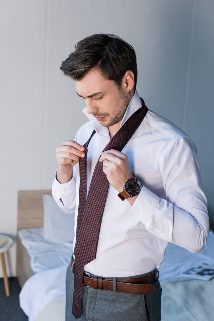 guapo confiado hombre poner corbata mientras de pie cerca de ropa de cama en casa
 - Foto, Imagen