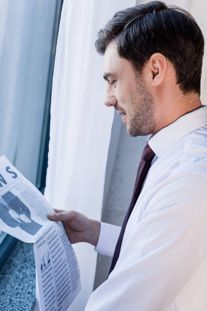 χαμογελαστός άντρας με λευκό πουκάμισο όρθιος στο παράθυρο και διαβάζοντας εφημερίδα - Φωτογραφία, εικόνα