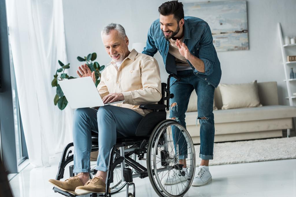 cheerful disabled retired man sitting in wheelchair near bearded son waving hand while having video call on laptop - Photo, Image