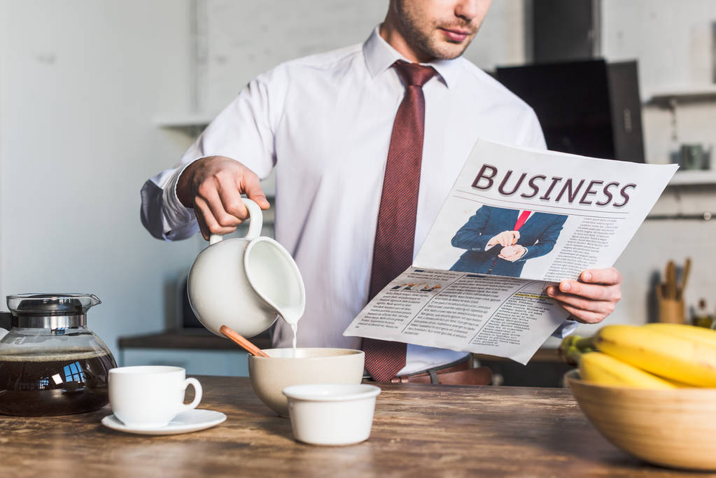 обрізаний вид чоловіка, який читає бізнес-газету, стоячи за кухонним столом і заливаючи молоко в миску
 - Фото, зображення