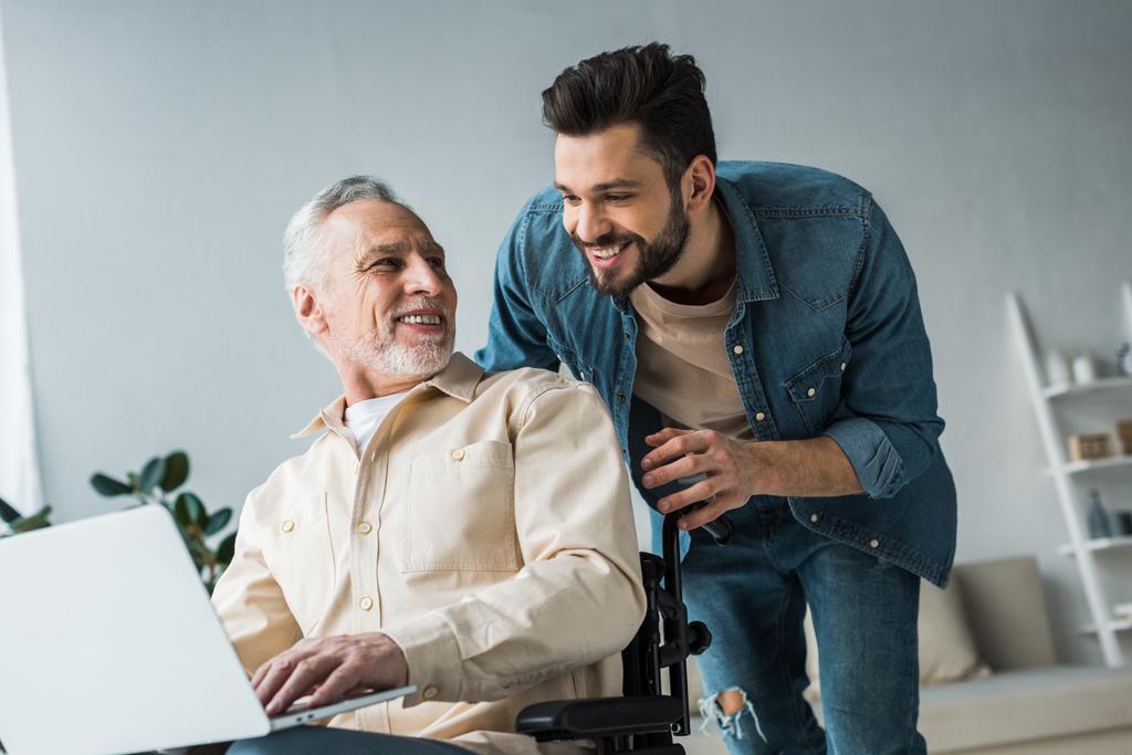 веселый пенсионер-инвалид сидит в инвалидной коляске рядом с красивым сыном и смотрит на ноутбук
 - Фото, изображение