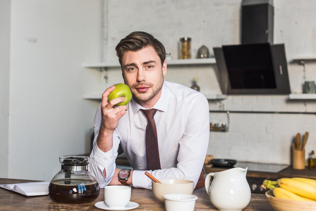 gutaussehender Mann mit Apfel und Blick in die Kamera, während er zu Hause in der Küche steht - Foto, Bild