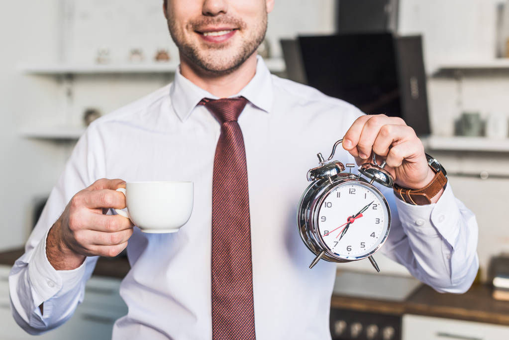 cropped view of man holding coffee cup and alarm clock while standing in kitchen at home - Photo, Image