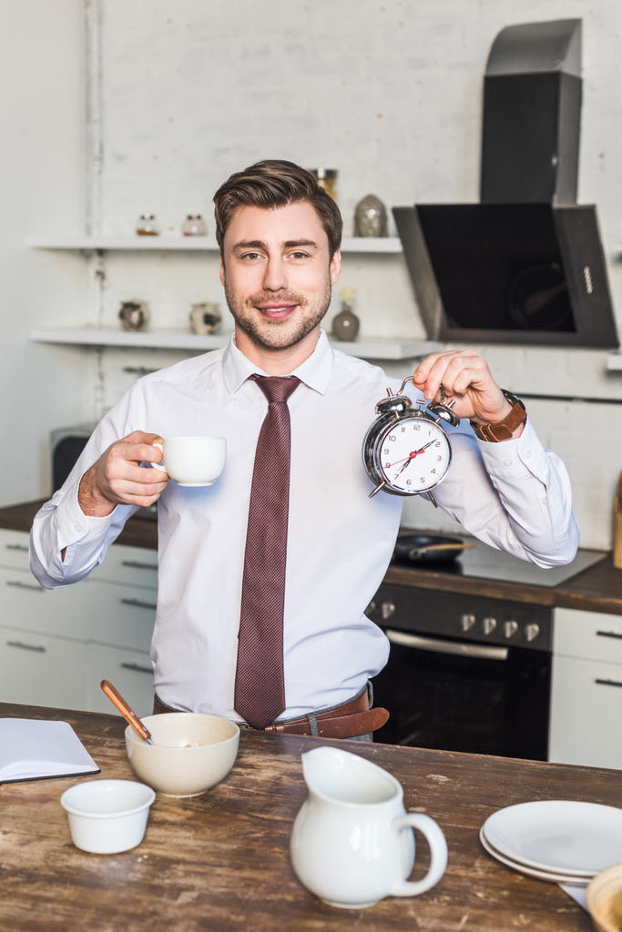 自宅でキッチンに立っている間、コーヒーカップと目覚まし時計を持つ陽気な男 - 写真・画像