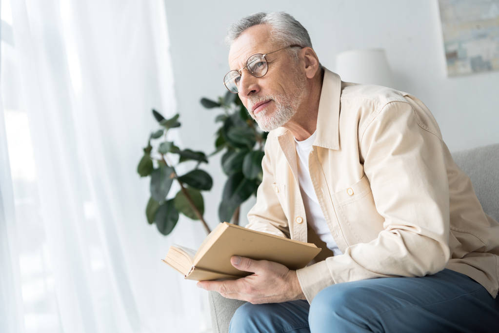 uomo anziano in bicchieri tenendo libro mentre seduto a casa
  - Foto, immagini