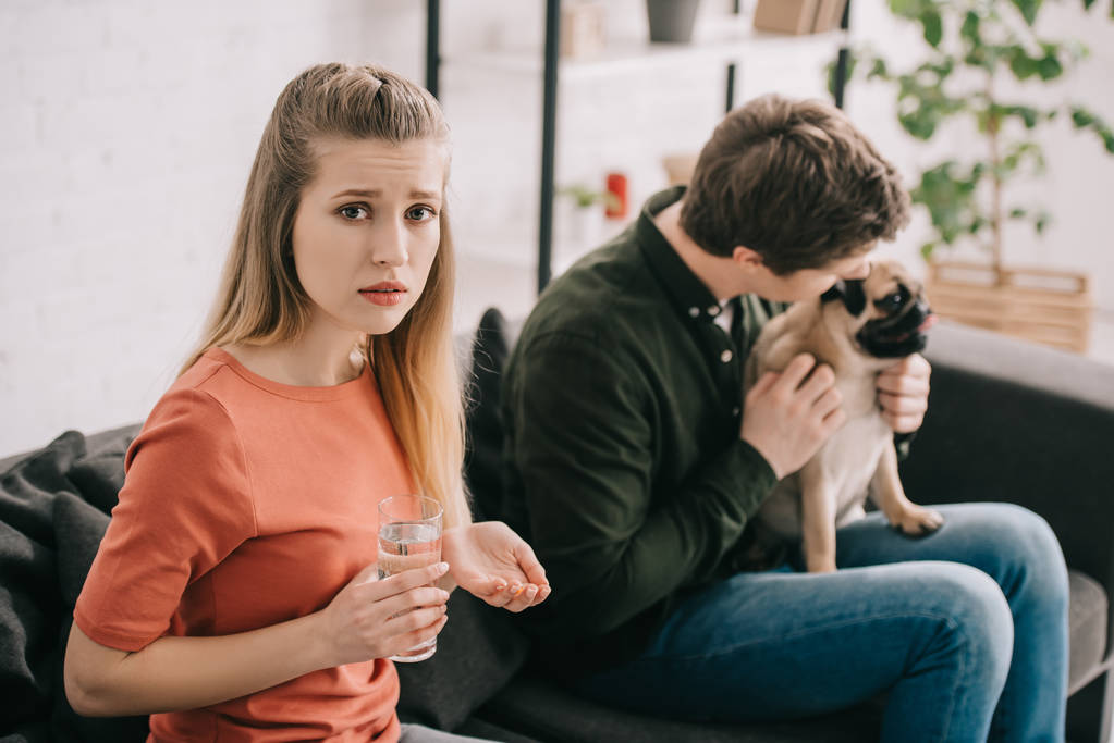 селективный фокус расстроенной женщины с аллергией на собаку с таблетками и стаканом воды рядом с мужчиной с мопсом
   - Фото, изображение