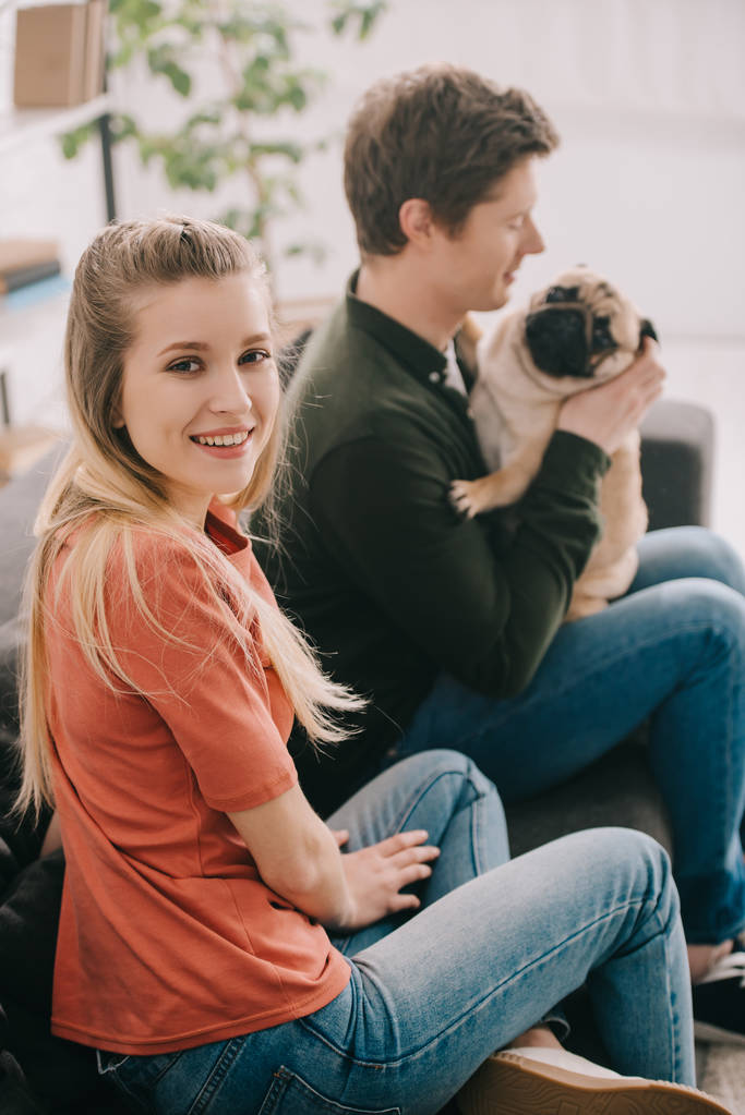 Selektywny fokus szczęśliwa Kobieta blonde, patrząc na kamery w pobliżu szczęśliwym człowiekiem z słodkie Mops pies  - Zdjęcie, obraz