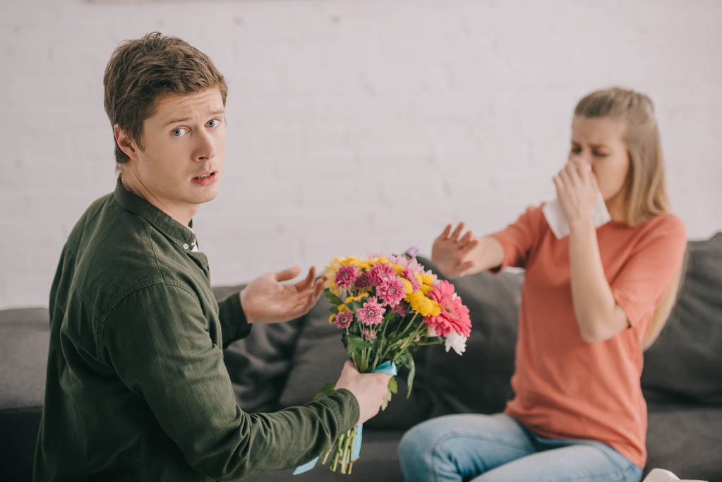 upset man holding flowers and looking at camera near blonde woman with pollen allergy sneezing in tissue  - Photo, Image