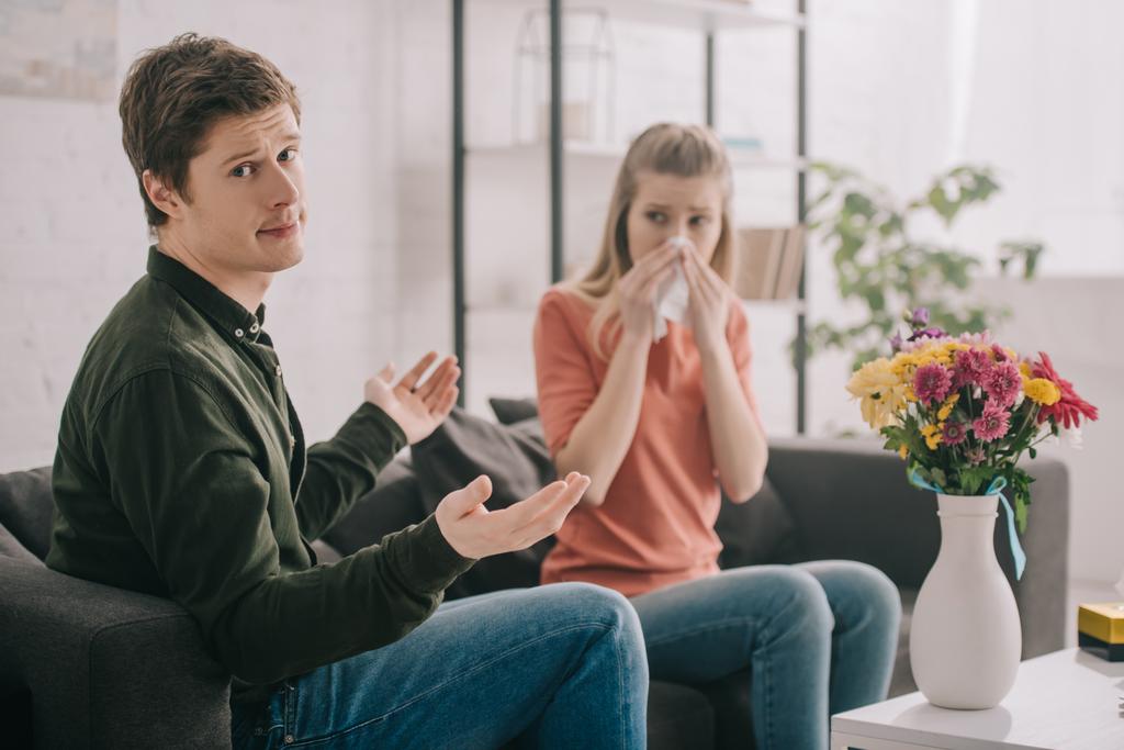 handsome man showing shrug gesture while sitting near vase with flowers and sneezing girl with pollen allergy - Photo, Image