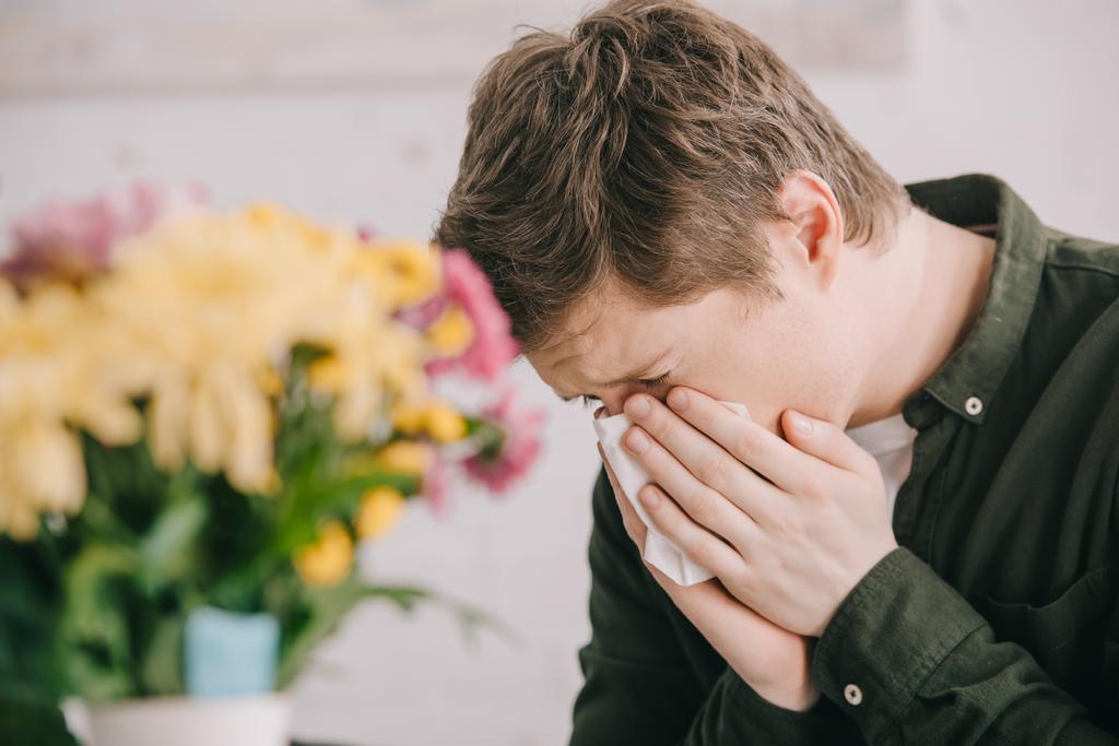 επιλεκτική εστίαση του φτερνίσματος ανθρώπου με αλλεργία στη γύρη κοντά ανθισμένα λουλούδια  - Φωτογραφία, εικόνα