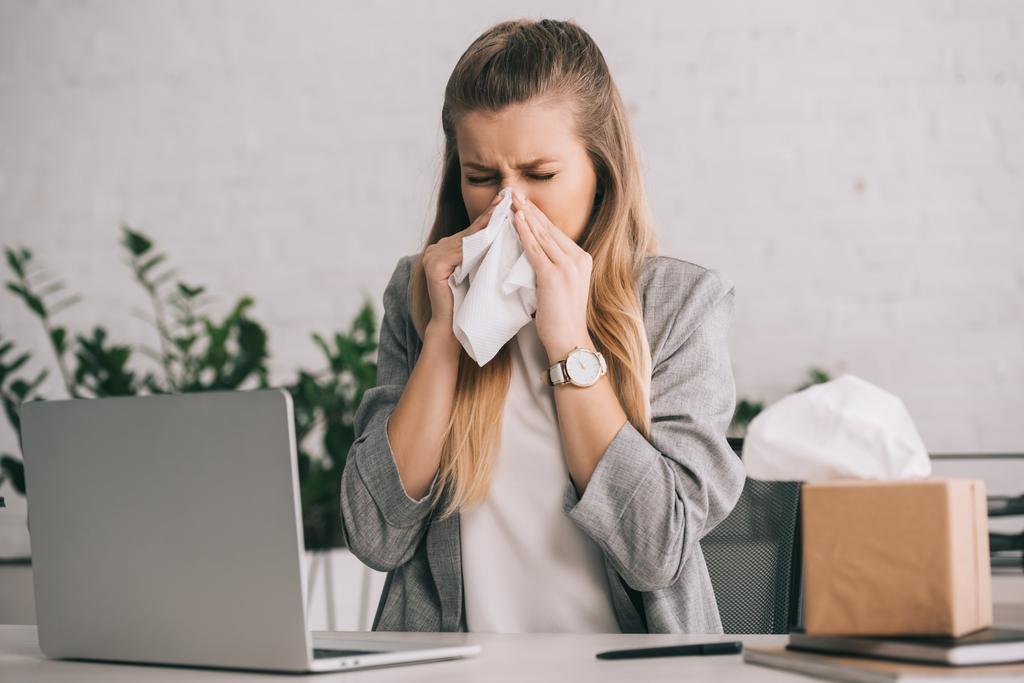 mujer de negocios rubia estornudando en tejido cerca de la computadora portátil en la oficina
  - Foto, Imagen