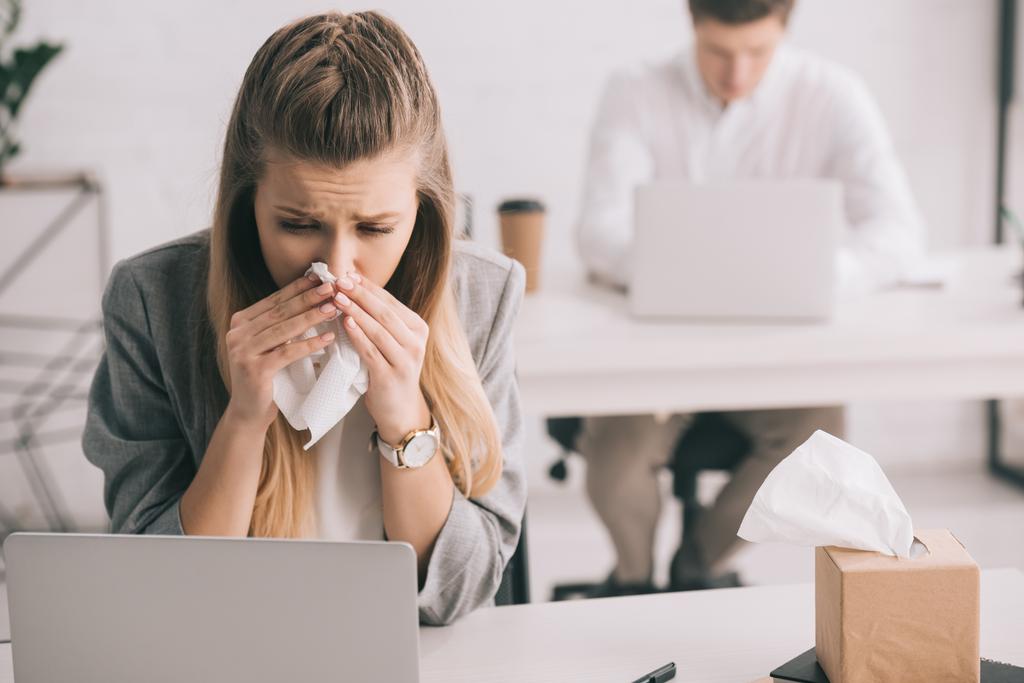 mulher de negócios loira espirrando em tecido perto de colega de trabalho no escritório
  - Foto, Imagem