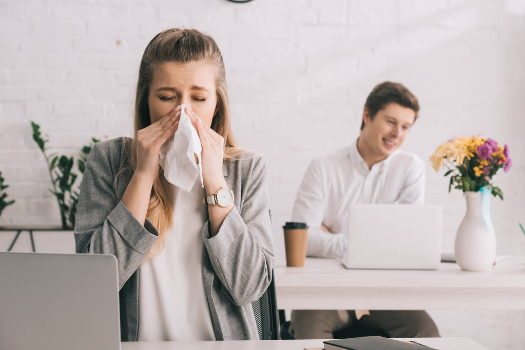 rubia mujer de negocios estornudando en tejido con los ojos cerrados cerca de compañero de trabajo en la oficina
  - Foto, imagen