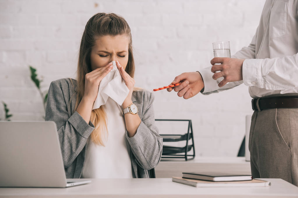 rubia empresaria estornudando en tejido con los ojos cerrados cerca de compañero de trabajo sosteniendo píldoras y vaso de agua
  - Foto, imagen