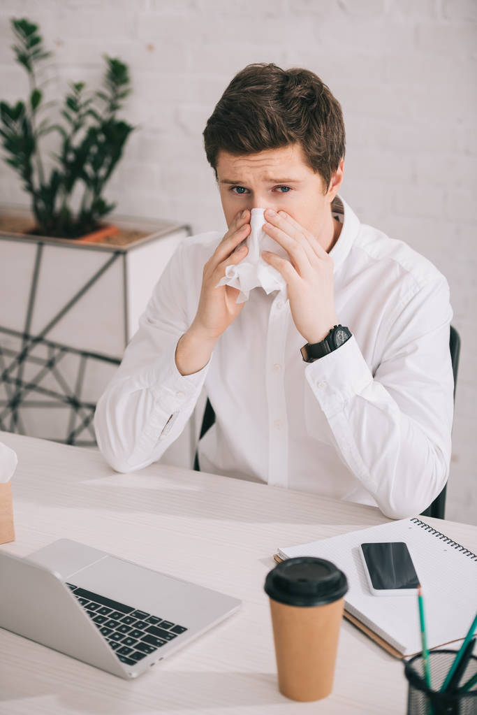 schöner Geschäftsmann niest in Taschentuch, während er neben Laptop und Smartphone im Büro sitzt  - Foto, Bild