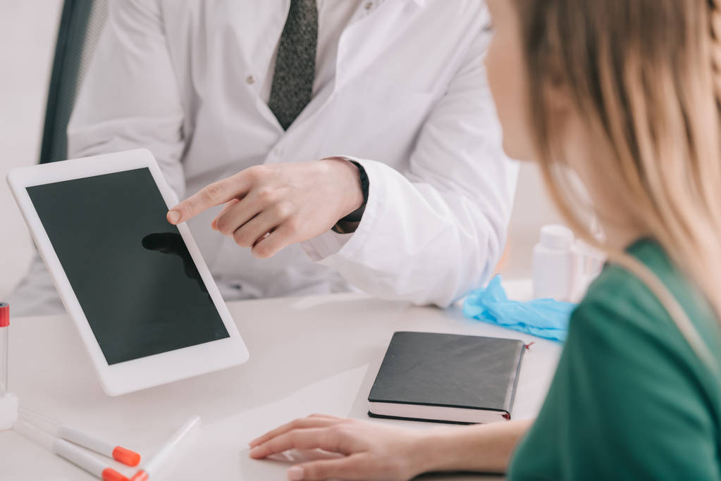金髪患者近く空白の画面とデジタル タブレットで指で指している白衣の医者のビューをトリミング  - 写真・画像