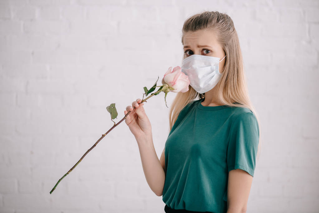 verängstigte Frau mit Pollenallergie trägt medizinische Maske und riecht Rose  - Foto, Bild