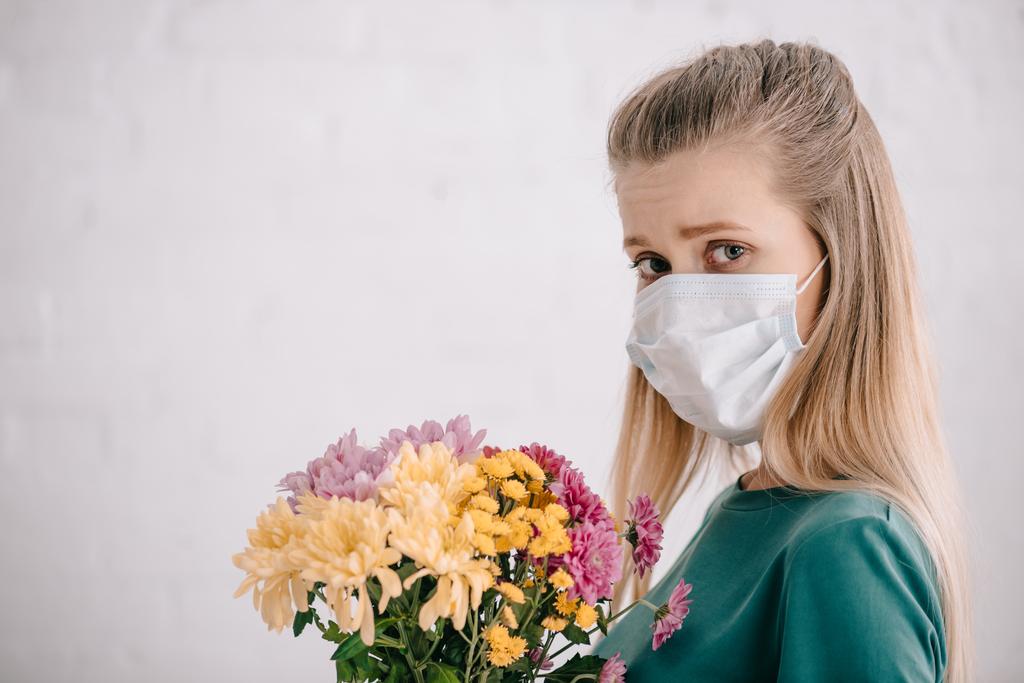 szőke nő, pollen allergia, orvosi maszkot visel, és a gazdaság virágok, miközben nézi a kamera  - Fotó, kép